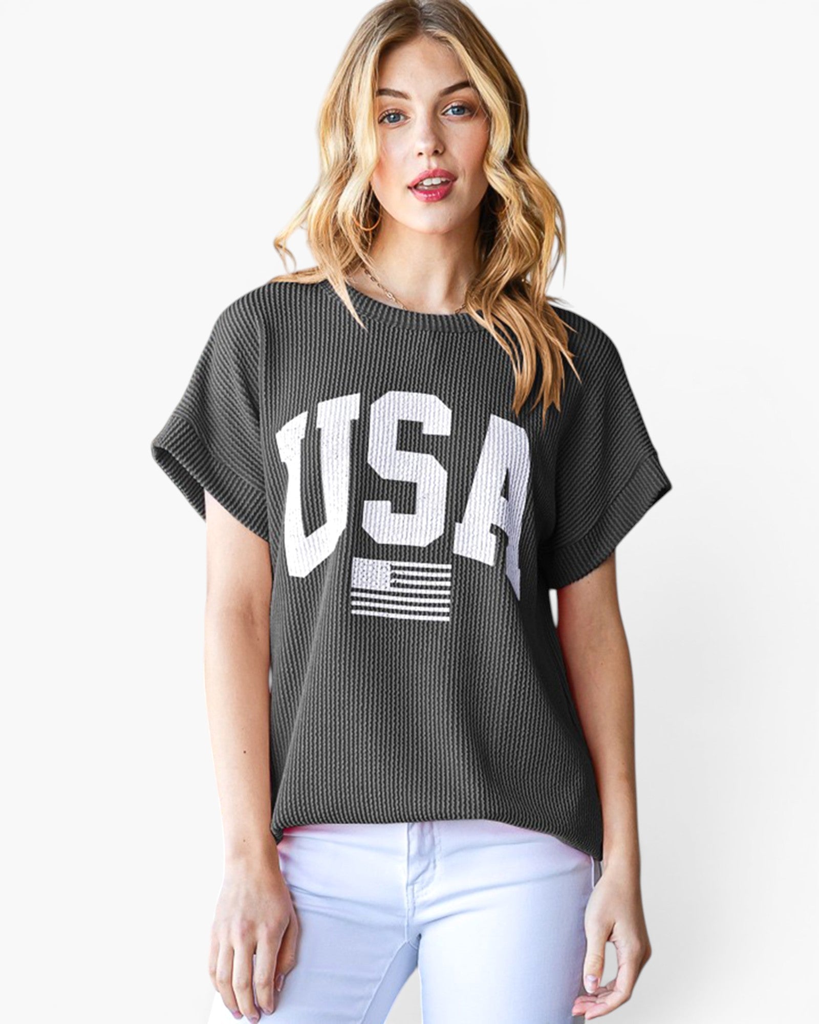 A woman wearing a dark gray ribbed short-sleeve t-shirt with a large white 'USA' and American flag graphic on the front, paired with white pants, against a plain white background.