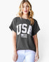 A woman wearing a dark gray ribbed short-sleeve t-shirt with a large white 'USA' and American flag graphic on the front, paired with white pants, against a plain white background.