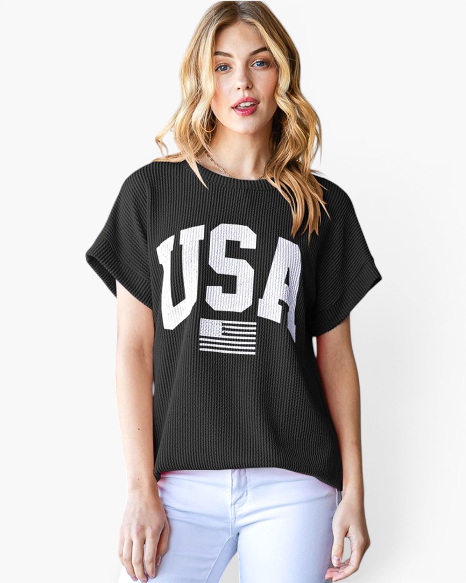 A woman wearing a black short-sleeve ribbed knit top with a large white 'USA' graphic and a small American flag on the front, paired with white pants, against a light gray studio background.