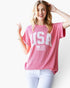 A woman wearing a pink short-sleeve ribbed t-shirt with a white 'USA' and American flag graphic on the front, paired with light blue jeans, against a plain white background.