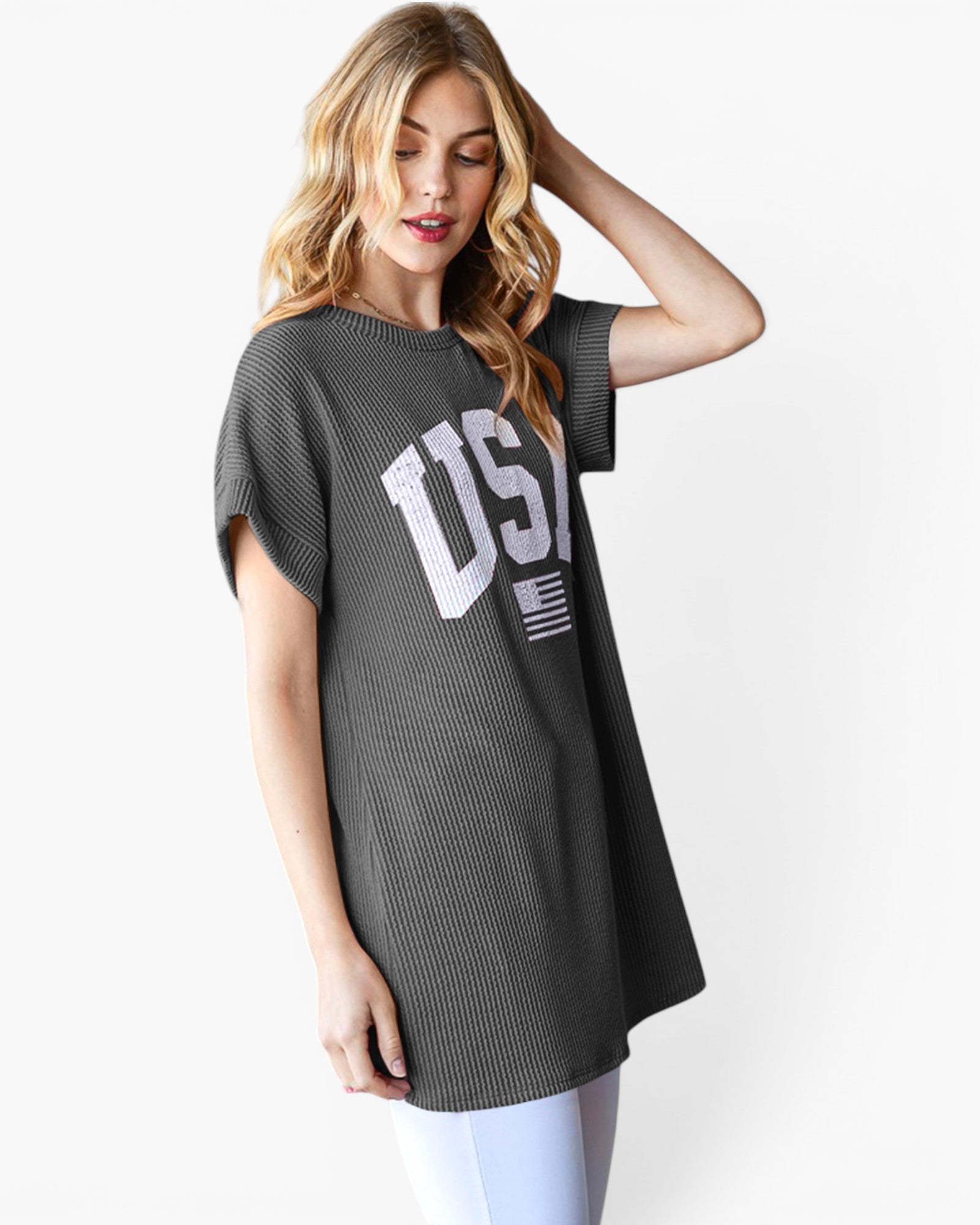 A woman wearing a dark gray ribbed short-sleeve tunic top with a large white 'USA' graphic and a small American flag on the front, paired with white leggings, against a light gray studio background.