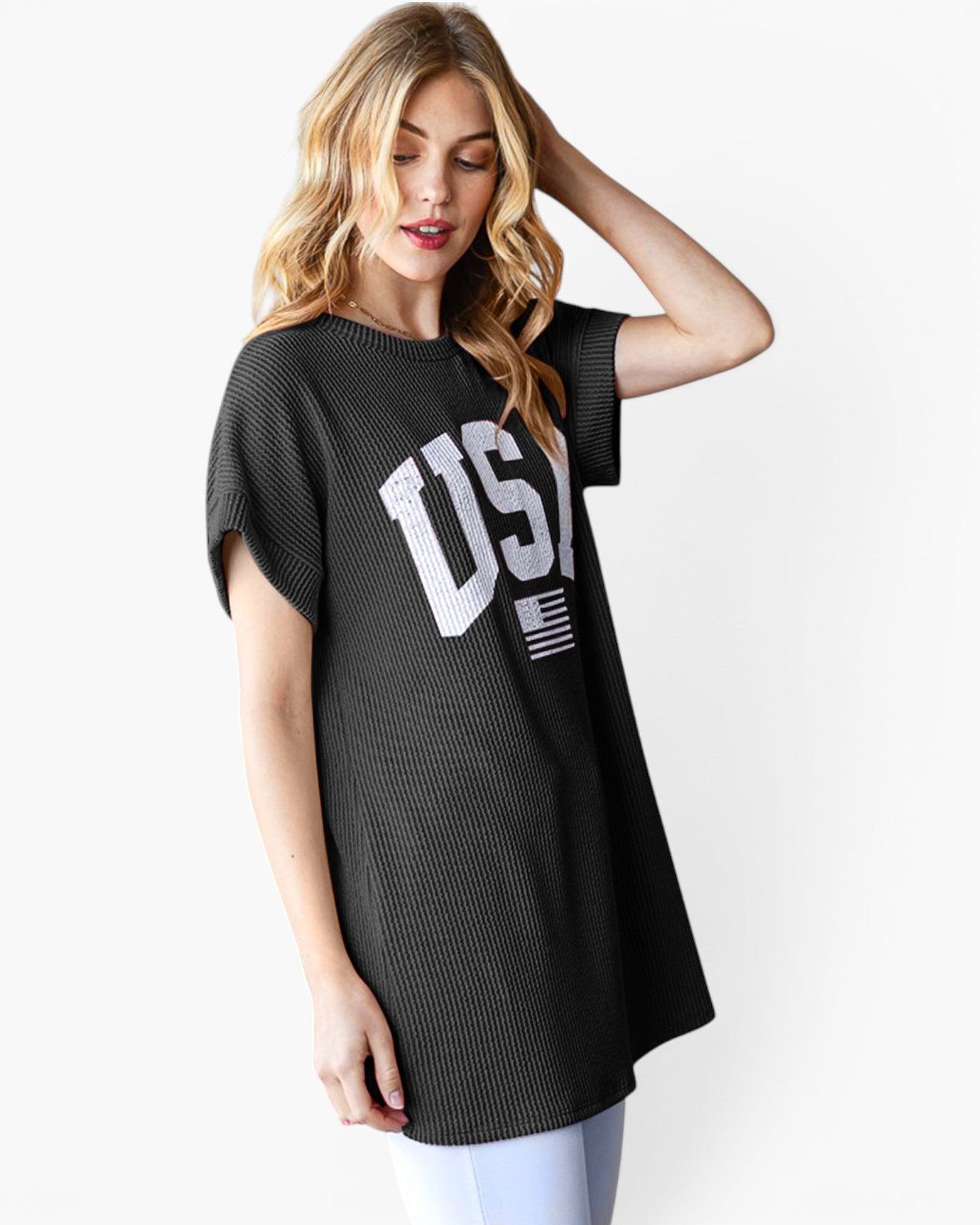 A woman wearing a black ribbed short-sleeve tunic top with a large white 'USA' and American flag graphic on the front, paired with light blue jeans, against a plain white background.