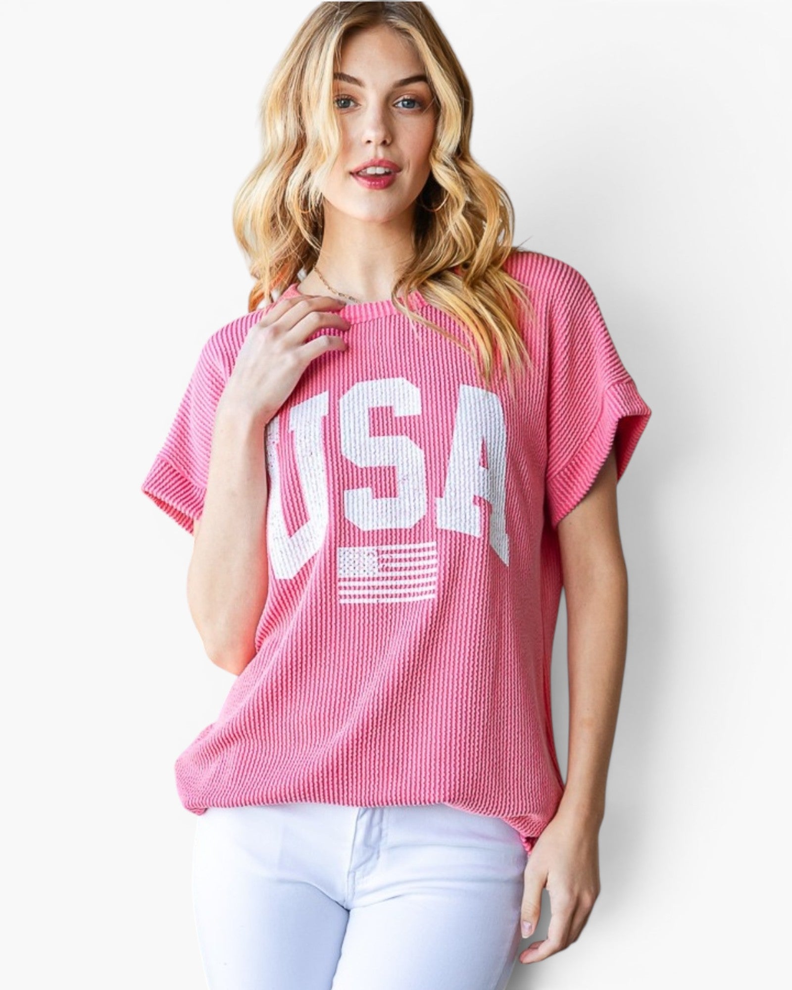 A woman wearing a pink short-sleeve waffle-knit top with a large white 'USA' and American flag graphic on the front, paired with light blue jeans, against a plain white background.