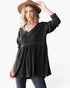 A woman wearing a black long-sleeve V-neck tunic top with a peplum waist, paired with dark jeans and a brown wide-brim hat, posing against a white studio background.