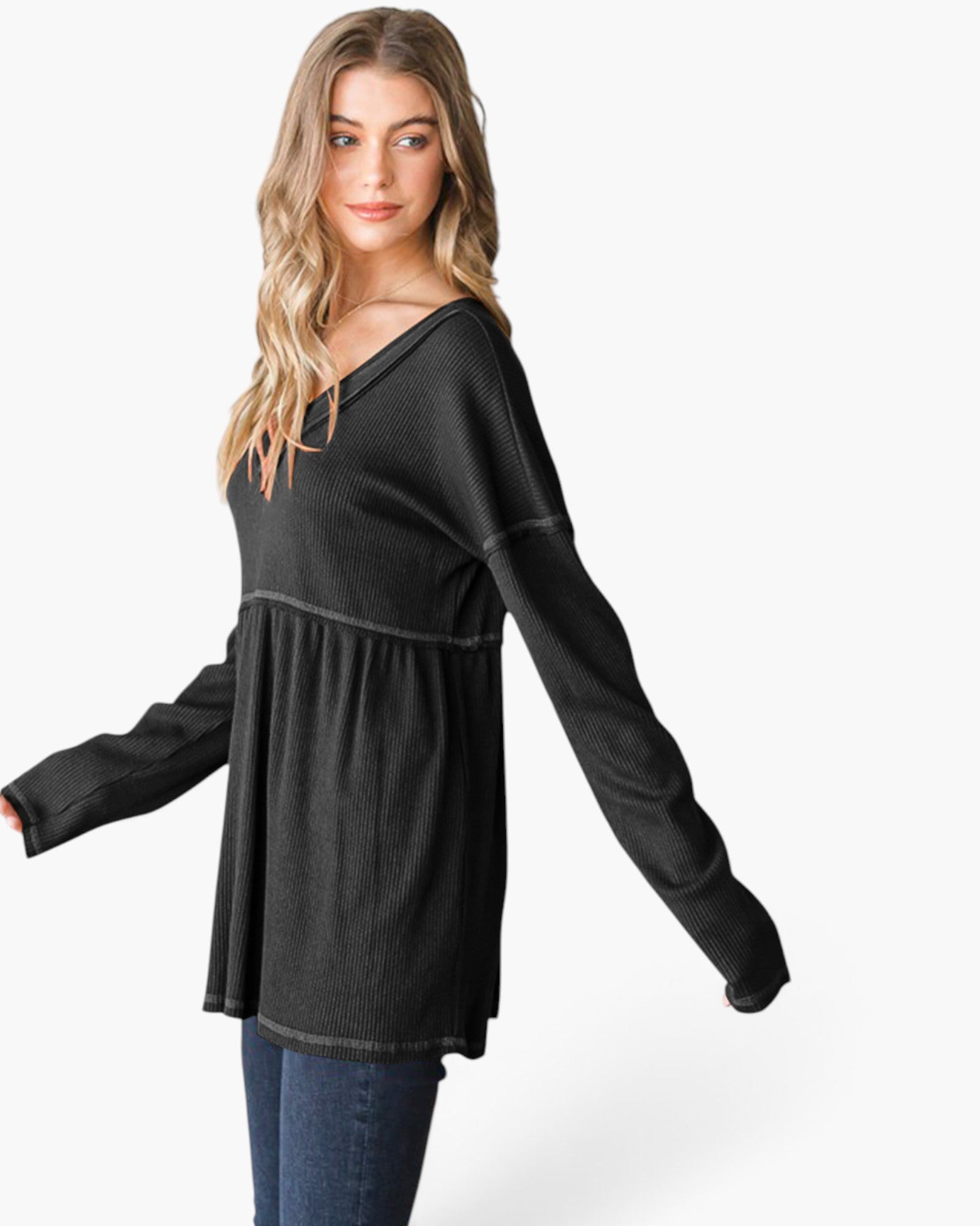 A woman modeling a black long-sleeve tunic top with a wide V-neck and gathered waist seam, paired with blue jeans against a light gray studio background.