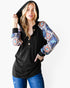 A woman modeling a black hooded long-sleeve top with patterned sleeves featuring a blue, orange, and white geometric Southwestern design, paired with blue jeans against a light gray background.