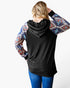 A woman shown from the back wearing a black long-sleeve hoodie with colorful geometric patterned sleeves, paired with blue jeans, against a plain white background.