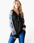A woman modeling a black long-sleeve henley top with colorful geometric patterned sleeves, paired with blue jeans, against a plain white studio background.