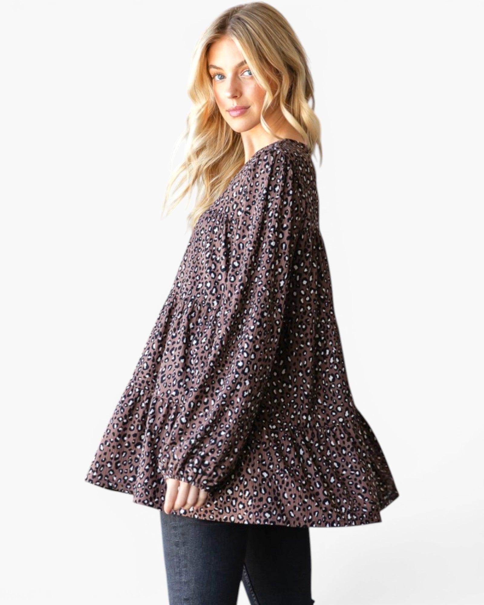 A woman wearing a dark brown long-sleeve tiered blouse with a small white floral pattern, paired with dark jeans, photographed against a plain white studio background.