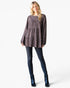 A woman modeling a long-sleeve, gathered peplum-style top with a dark brown and black speckled pattern, paired with dark blue skinny jeans and metallic blue ankle boots, against a plain white studio background.