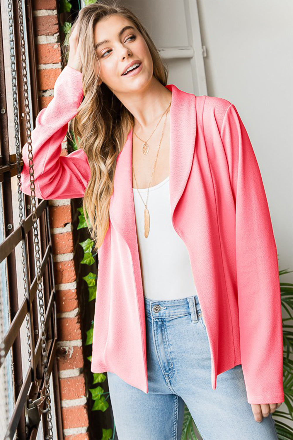 Woman wearing a pink jacket, white top, and blue jeans standing next to a brick wall.