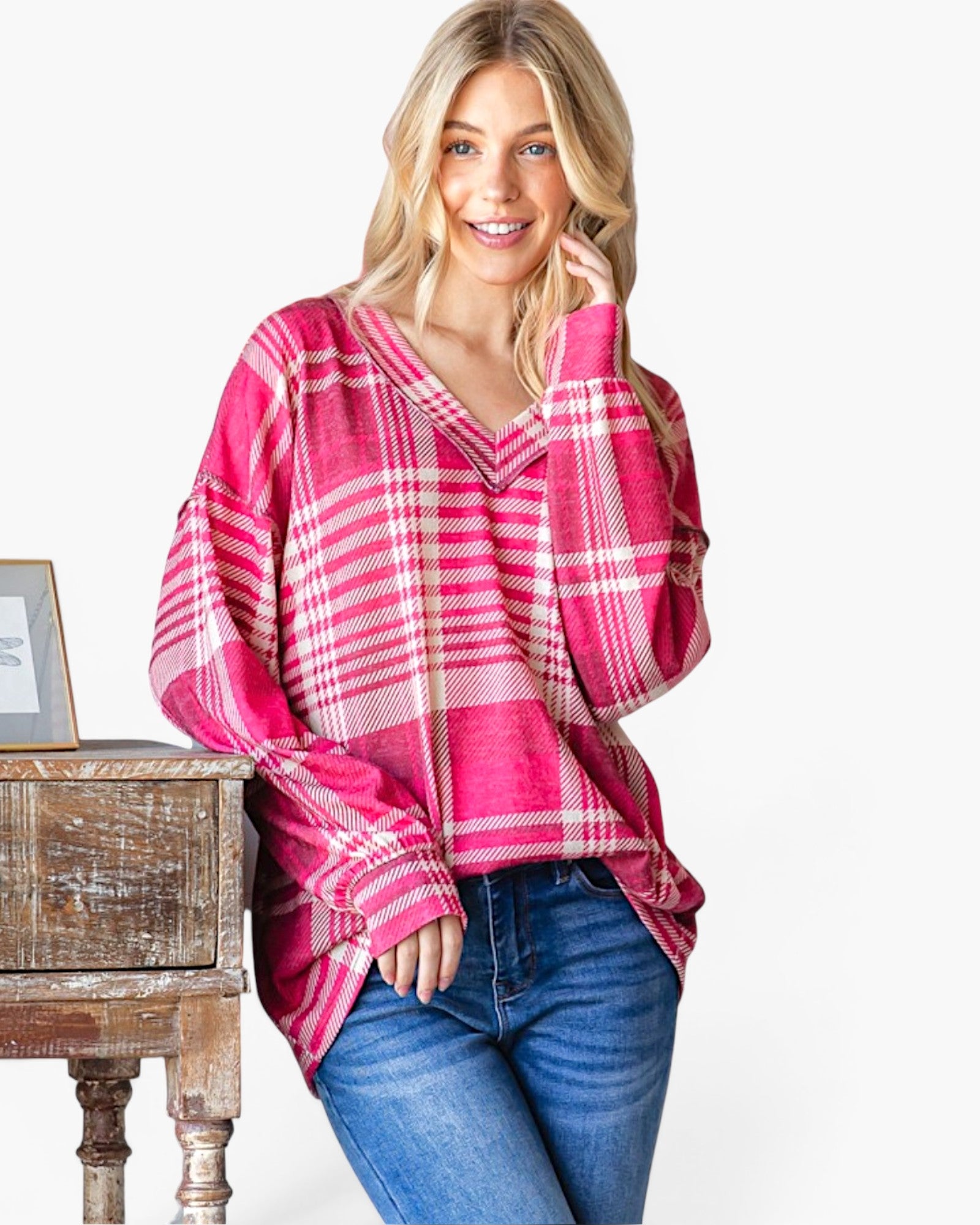 A woman wearing a pink and white plaid long-sleeve top with blue jeans, leaning against a rustic wooden side table against a plain white background.
