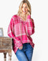 A woman wearing a pink and white plaid long-sleeve top with blue jeans, leaning against a rustic wooden side table against a plain white background.
