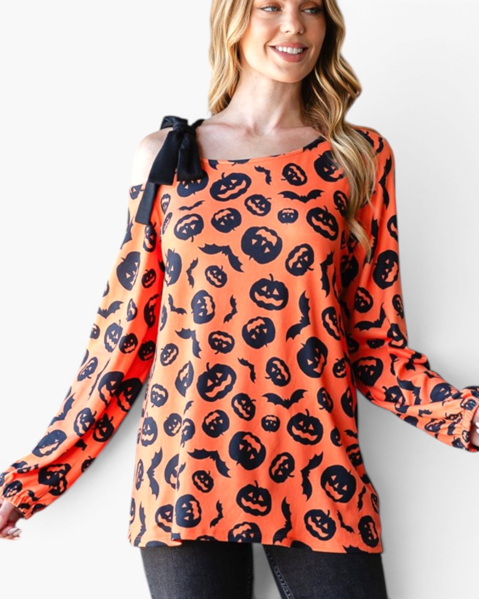 A woman wearing a long-sleeve orange top with a black pumpkin and bat print, featuring a black bow detail on the shoulder, set against a plain white background.