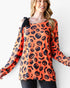 A woman wearing a long-sleeve orange top with a black pumpkin and bat print, featuring a black bow detail on the shoulder, set against a plain white background.