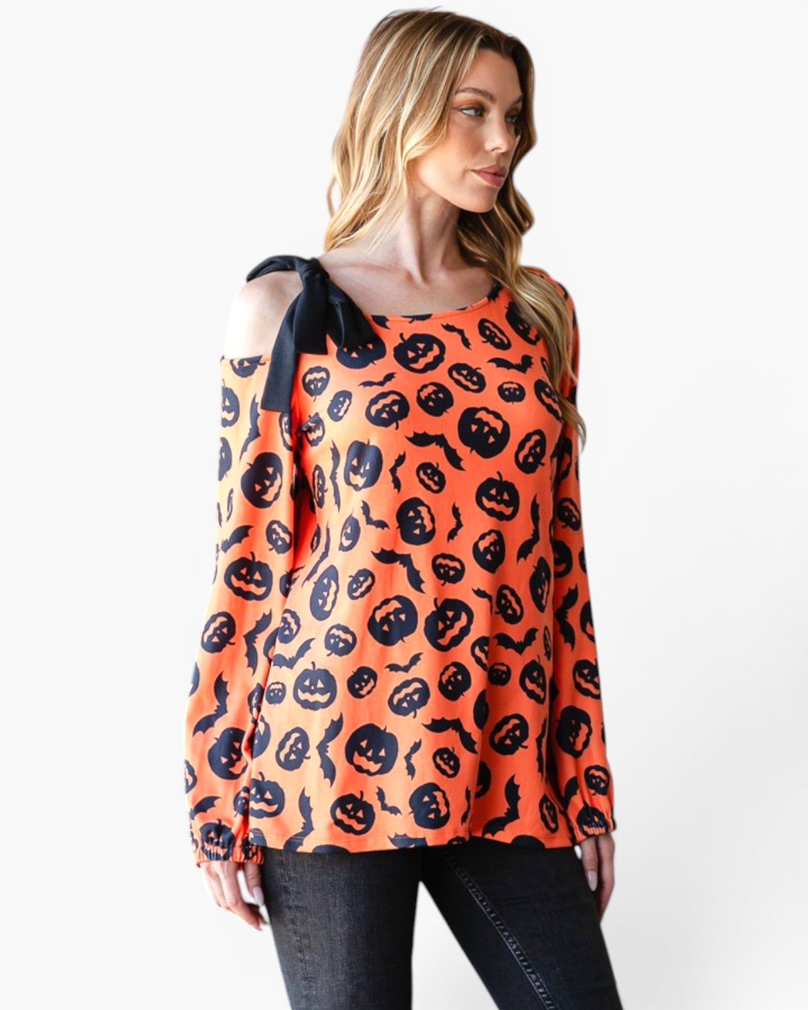 A woman wearing a long-sleeve orange top with a black pumpkin and bat pattern, featuring a cold-shoulder design with a black ribbon tie on the left shoulder, paired with dark jeans against a plain white background.