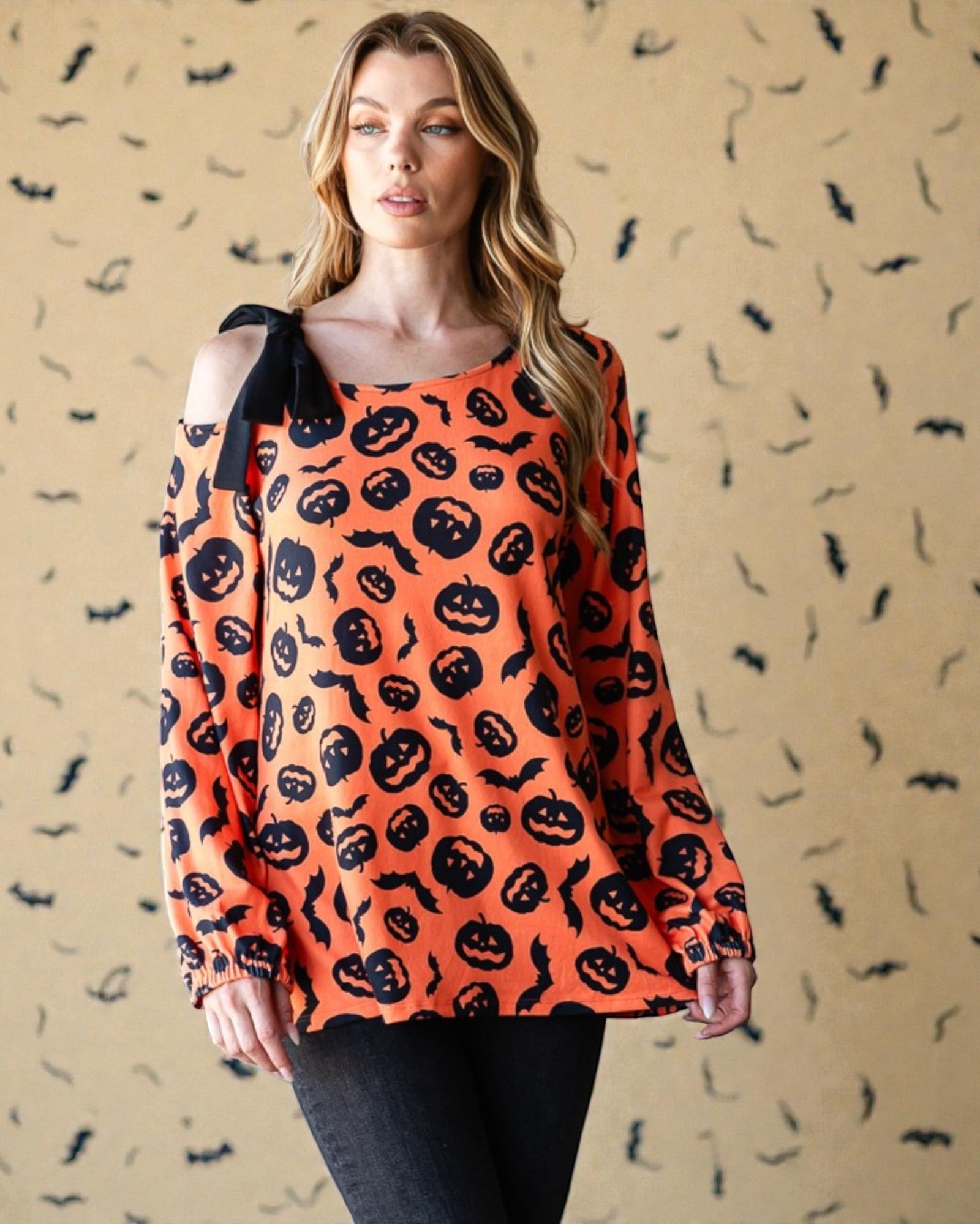Woman wearing an orange Halloween-themed long-sleeve top with black pumpkins and bats on a beige background.