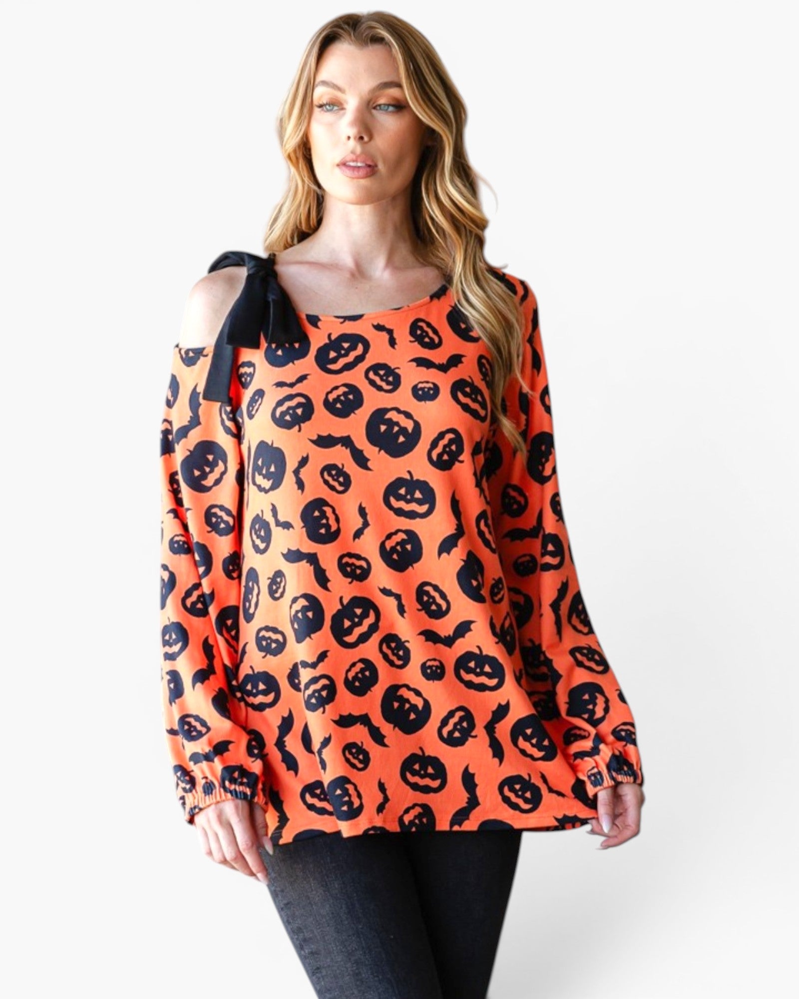 A woman modeling a long-sleeve orange top with a black pumpkin and bat pattern, featuring a black shoulder tie detail, paired with dark jeans against a plain white background.