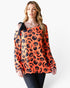 A woman modeling a long-sleeve orange top with a black pumpkin and bat pattern, featuring a black shoulder tie detail, paired with dark jeans against a plain white background.