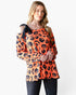 A woman modeling a long-sleeve orange top with a black pumpkin and bat print, featuring a black shoulder tie detail, paired with dark jeans against a plain white background.