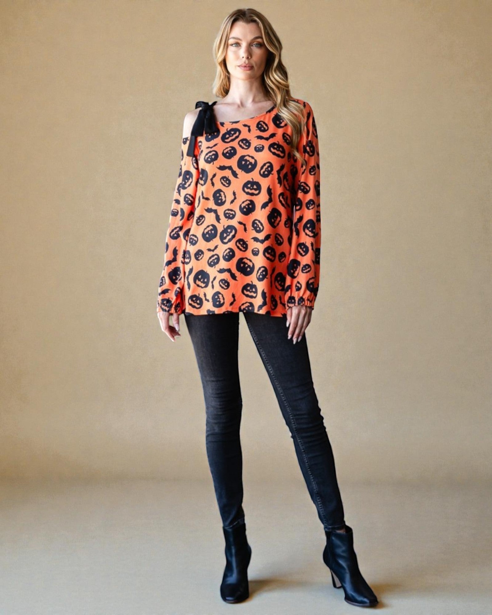 Woman wearing an orange sweater with black pumpkin pattern and black pants against a beige background