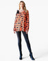 A woman modeling a long-sleeve orange top with a black and dark blue pumpkin print, paired with dark skinny jeans and blue heeled boots, against a plain white background.