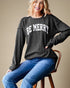 Woman wearing a black sweater with 'BE MERRY' text, sitting on a wooden stool against a beige background.