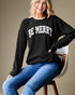 Woman wearing a black sweater with 'BE MERRY' text, sitting on a chair against a beige background
