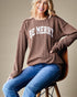 Woman wearing a brown sweater with 'BE MERRY' text, sitting on a chair against a beige background