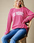 Woman wearing a pink sweater with 'BE MERRY' text, sitting on a wooden chair.