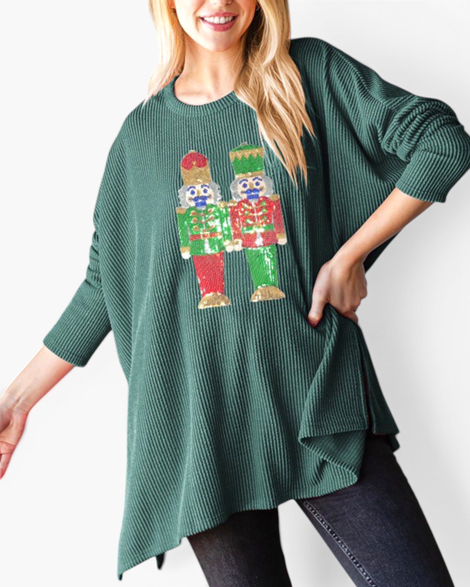 A woman wearing a dark green ribbed knit tunic sweater with a colorful sequined nutcracker graphic on the front, paired with dark jeans against a plain white background.