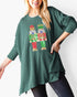 A woman wearing a dark green ribbed knit tunic sweater with a colorful sequined nutcracker graphic on the front, paired with dark jeans against a plain white background.