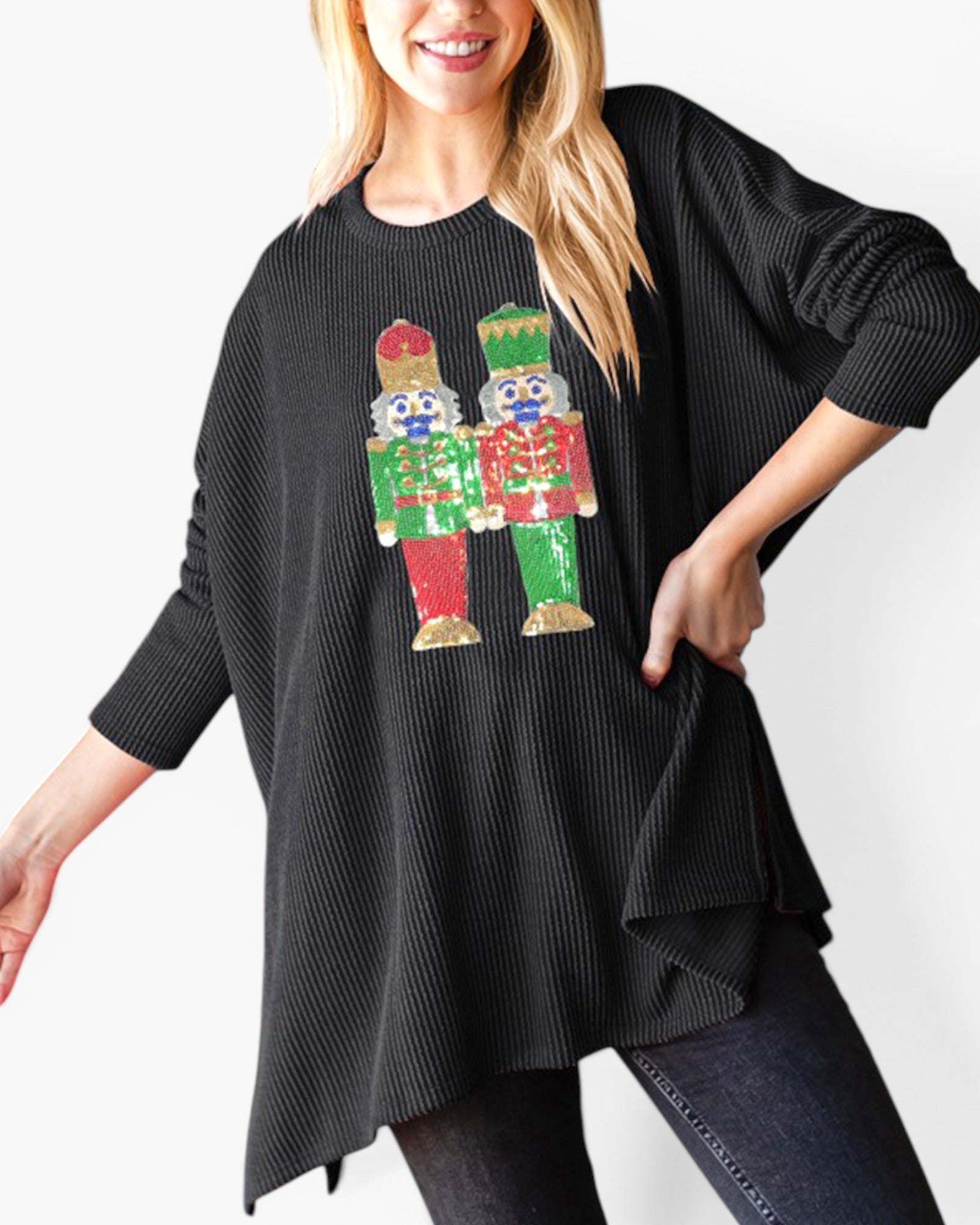 A woman wearing a black ribbed knit tunic sweater with a colorful sequined nutcracker graphic on the front, paired with dark jeans against a plain white background.