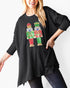 A woman wearing a black ribbed knit tunic sweater with a colorful sequined nutcracker graphic on the front, paired with dark jeans against a plain white background.