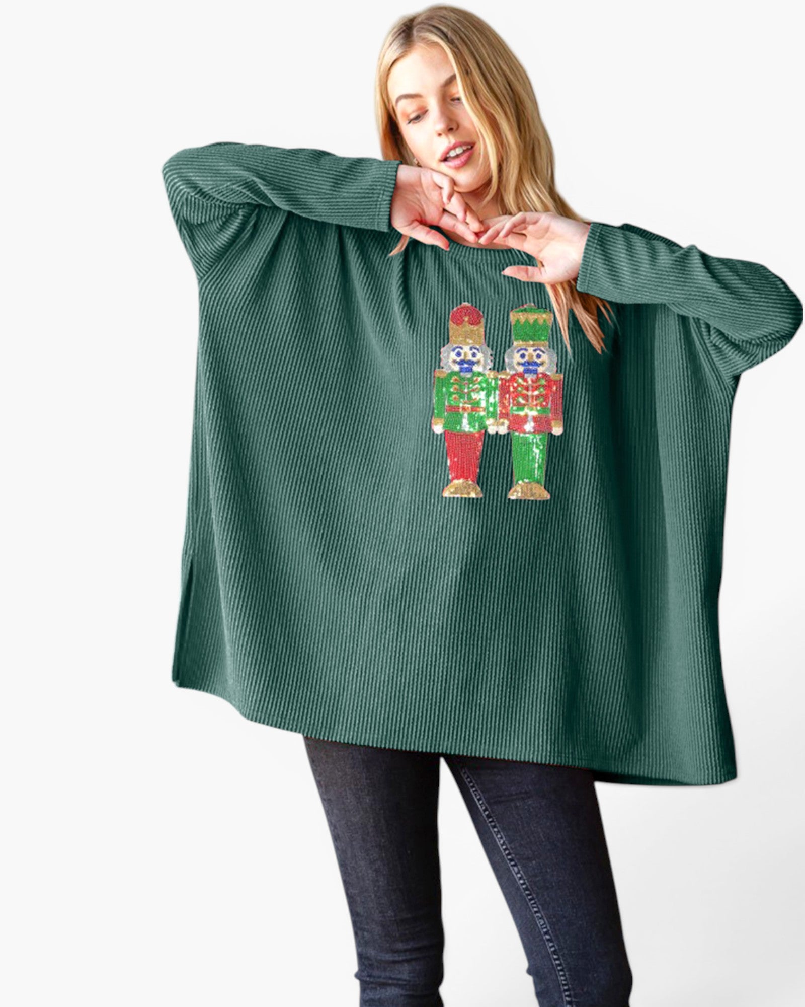 A woman modeling a dark green ribbed knit poncho-style sweater with a colorful nutcracker graphic on the front, paired with dark jeans against a plain white background.