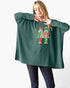 A woman modeling a dark green ribbed knit poncho-style sweater with a colorful nutcracker graphic on the front, paired with dark jeans against a plain white background.