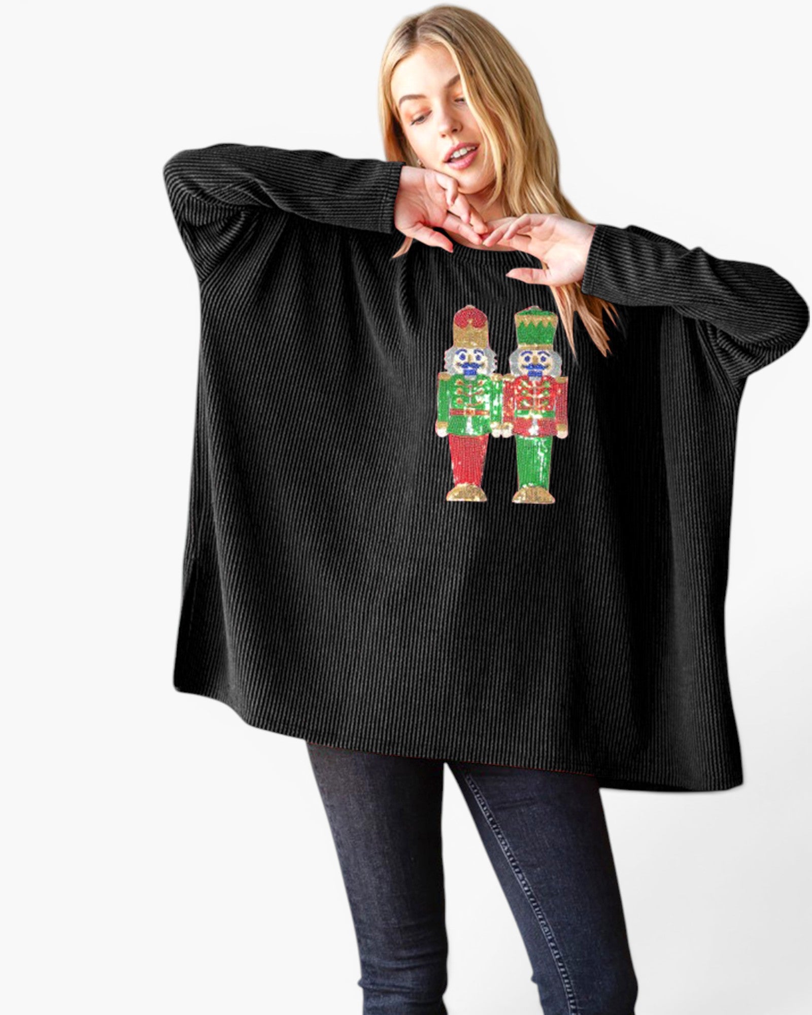 A woman modeling a black ribbed knit poncho sweater with a colorful nutcracker graphic on the front, paired with dark jeans against a plain white background.