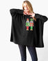 A woman modeling a black ribbed knit poncho sweater with a colorful nutcracker graphic on the front, paired with dark jeans against a plain white background.