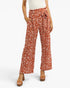 A model wearing high-waisted, wide-leg pants in a rust-orange color with a white leaf pattern, paired with a white top and brown platform sandals, against a light gray background.