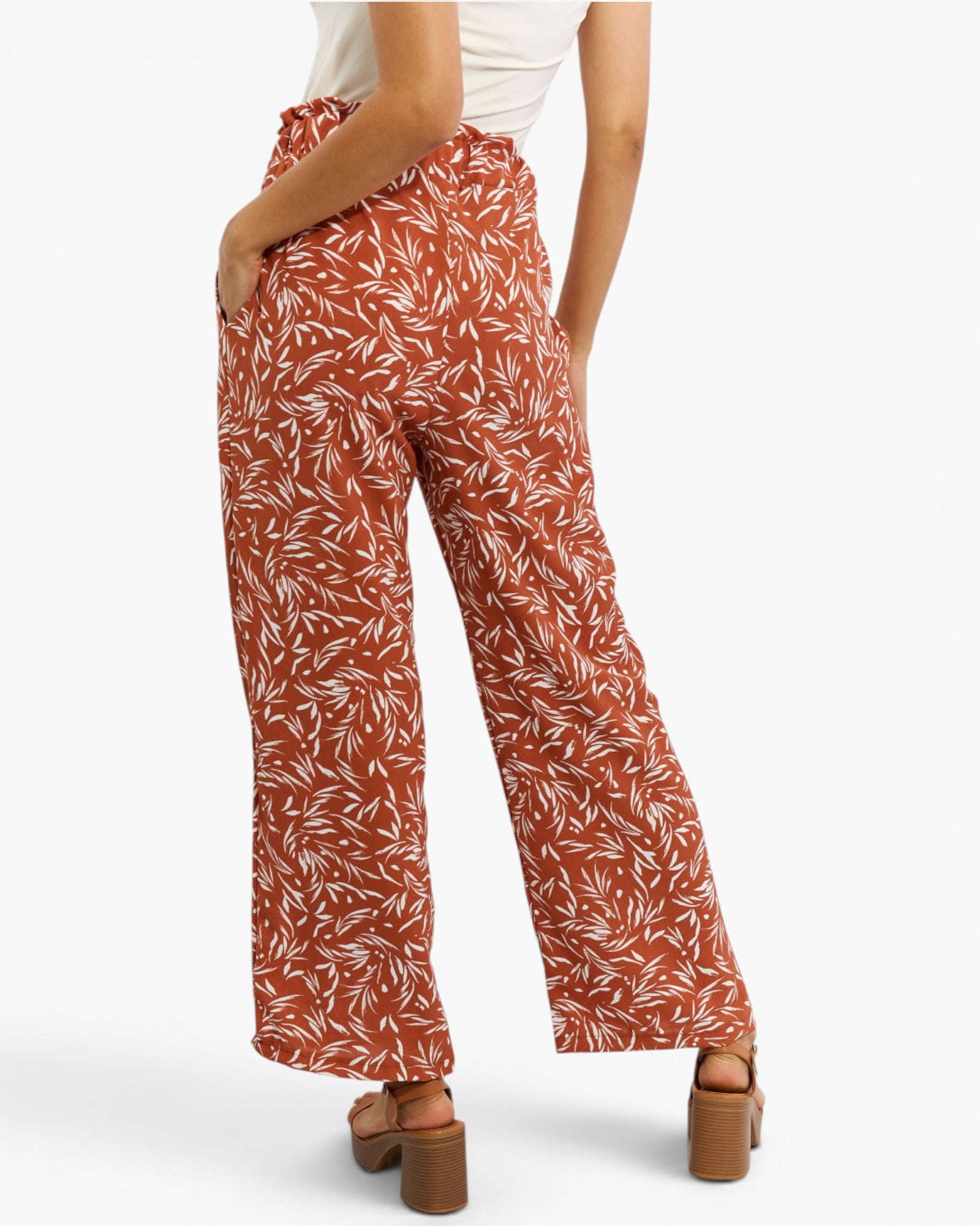 A person shown from the back wearing rust-orange wide-leg pants with a white leaf print, paired with a white top and brown block-heel sandals, against a light gray studio background.