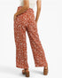 A person shown from the back wearing rust-orange wide-leg pants with a white leaf print, paired with a white top and brown block-heel sandals, against a light gray studio background.
