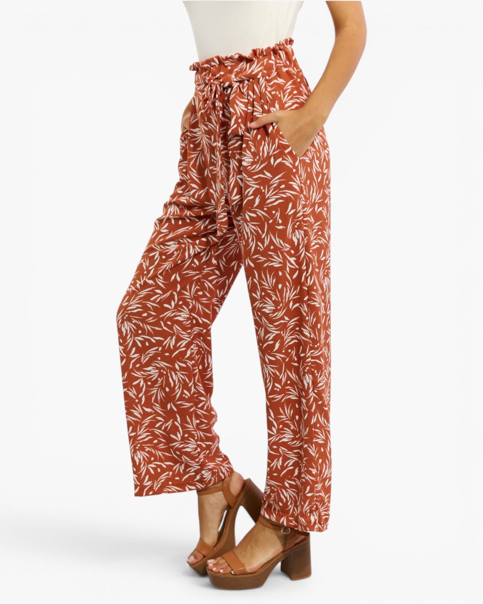 A model wearing high-waisted, wide-leg pants in a rust-orange and white leaf pattern, paired with brown platform sandals, shown from the waist down against a light gray background.