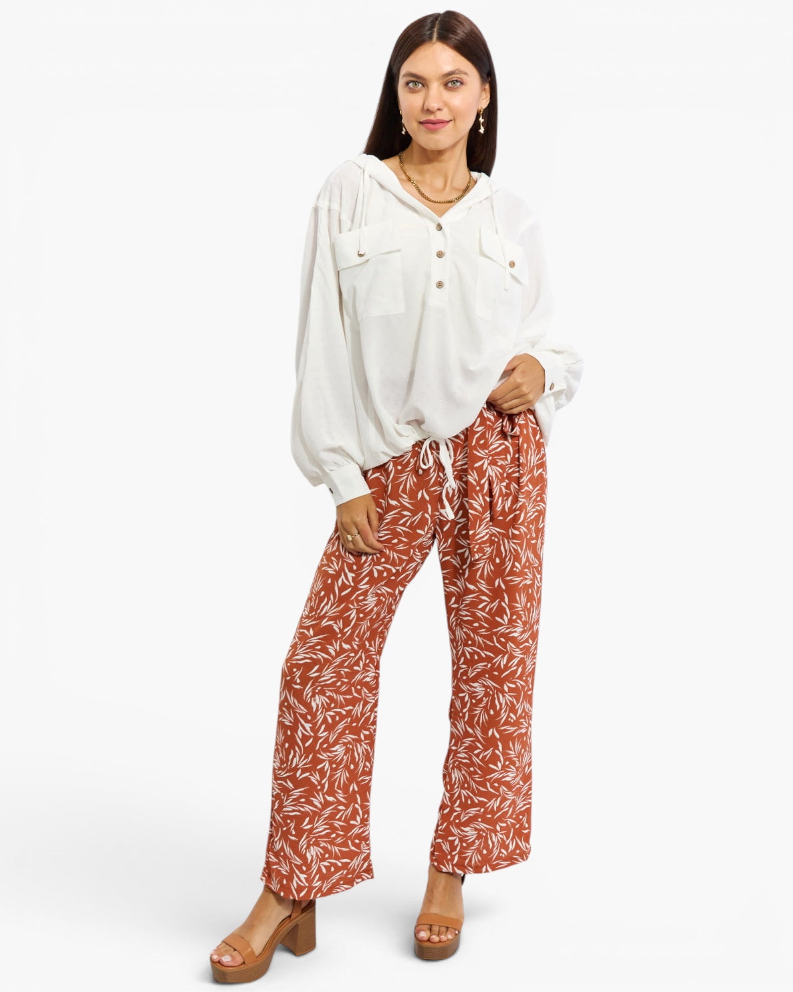 A woman modeling a white long-sleeve button-front blouse with a small tie detail at the collar, paired with rust-orange wide-leg pants featuring a white leafy pattern and brown platform sandals, against a plain white studio background.
