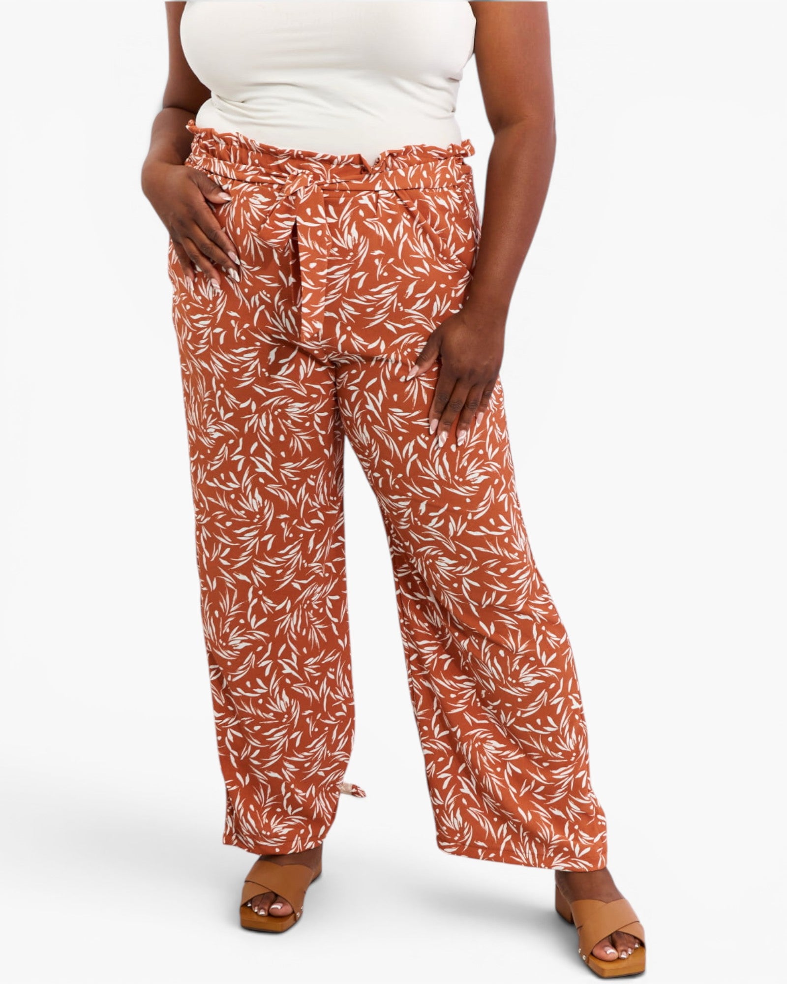 A model wearing a pair of wide-leg pants in rust orange with a white leaf pattern, paired with a white top and tan sandals, against a light gray background.