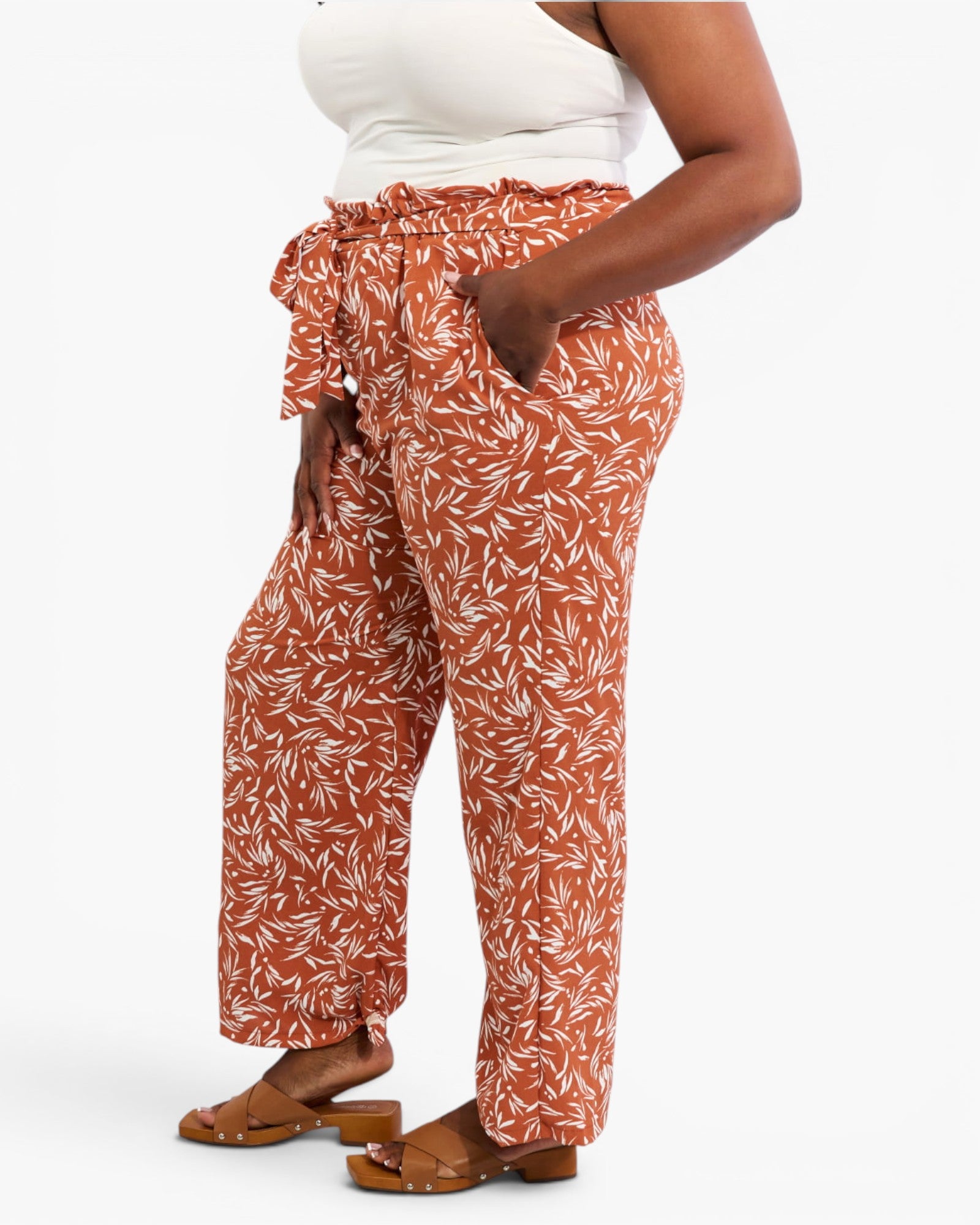 A model wearing high-waisted, wide-leg pants in a rust-orange color with a white leaf pattern, styled with a white tank top and brown sandals against a light gray background.