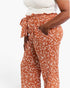 A close-up of a model wearing rust-orange wide-leg pants with a white leafy pattern, featuring a ruffled tie waist and side pockets.