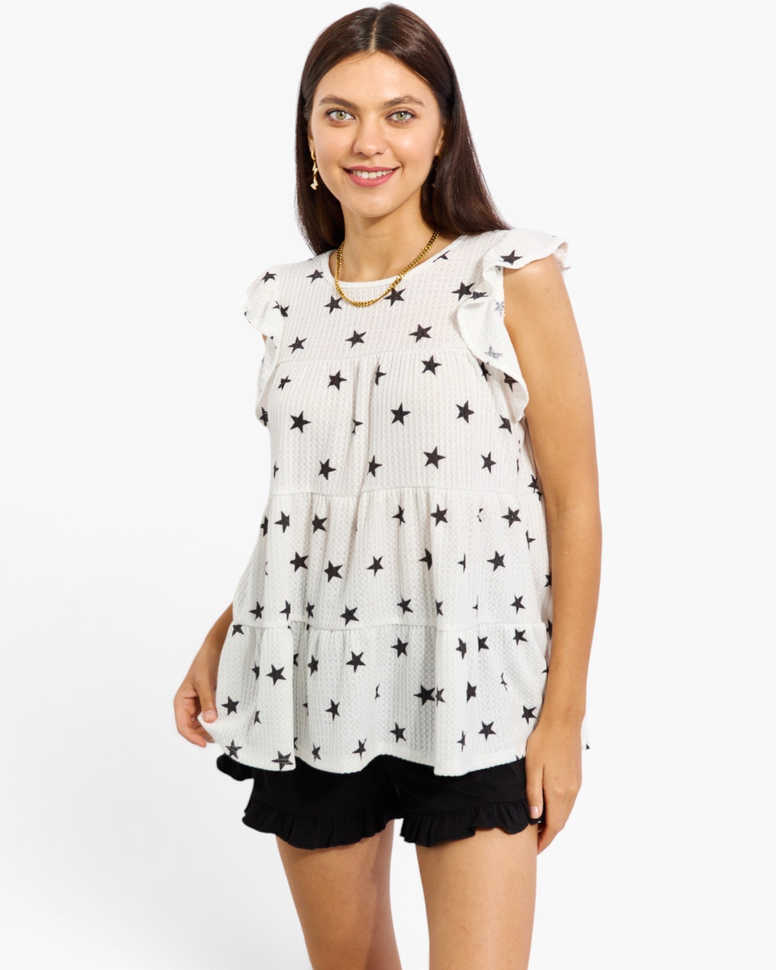A woman modeling a sleeveless white tiered top with a black star pattern and ruffled shoulder details, paired with black ruffled shorts, against a plain white studio background.