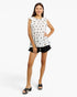 A woman modeling a sleeveless white top with a black star pattern and ruffled shoulders, paired with black shorts and black slide sandals, against a light gray studio background.