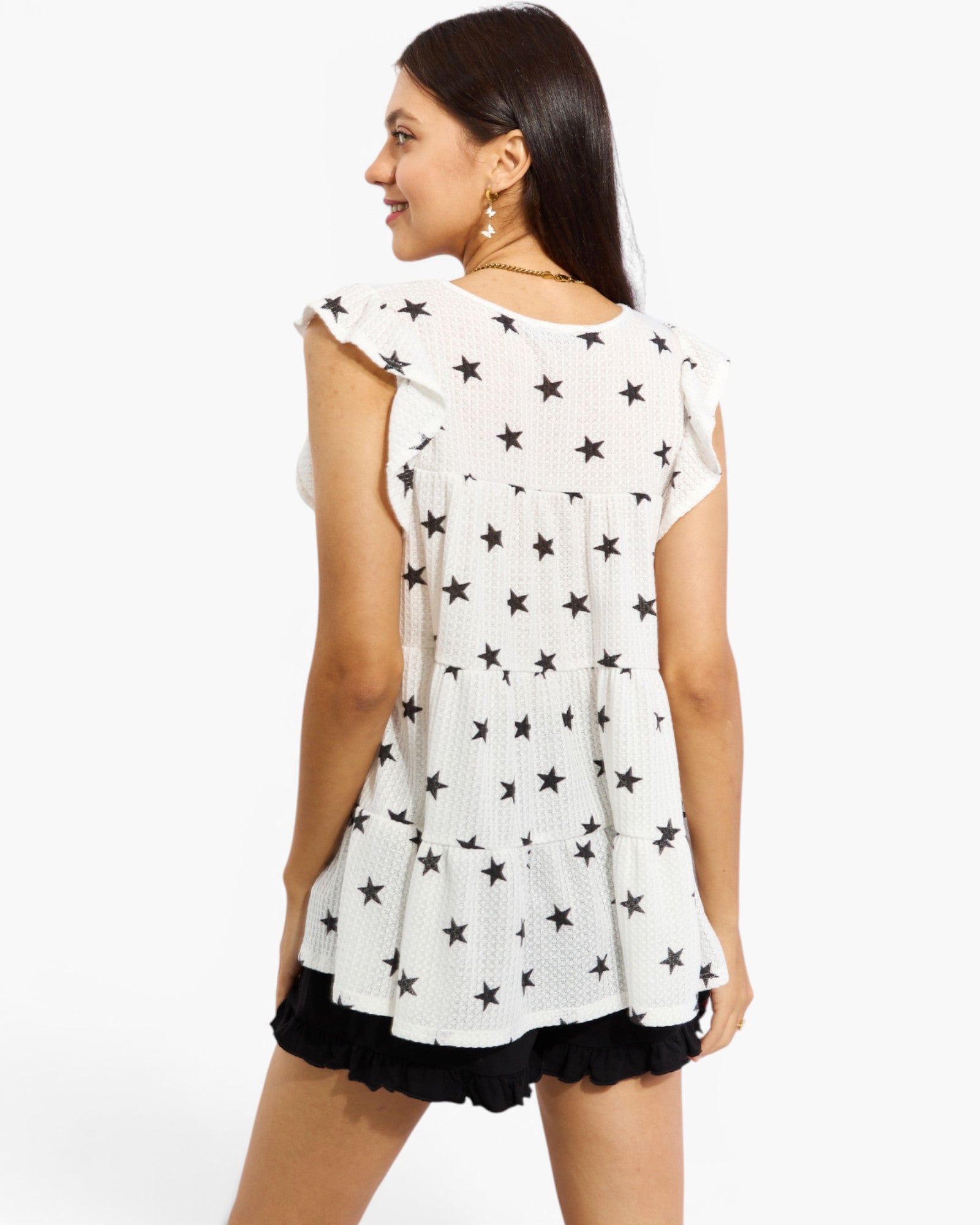 A woman shown from the back wearing a white sleeveless top with an allover black star pattern and black ruffled shorts, against a plain white studio background.