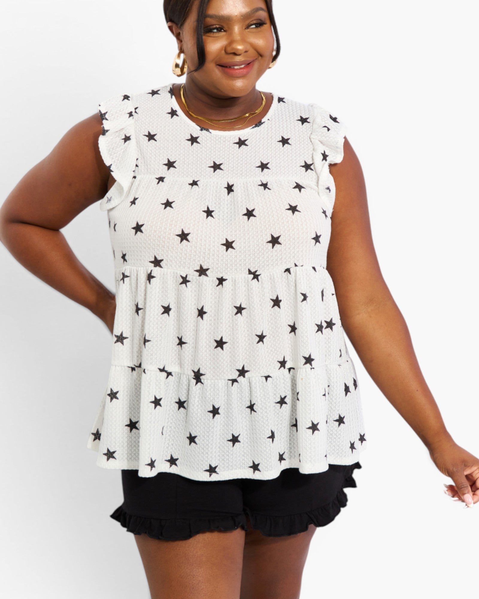 A woman wearing a white sleeveless ruffle-sleeve top with an all-over black star pattern and black ruffled shorts, photographed against a light gray studio background.
